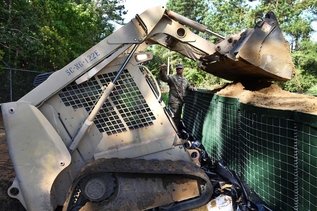 A soldier gives directions to another soldier operating a skid steer loader providing sand for flood water barriers.