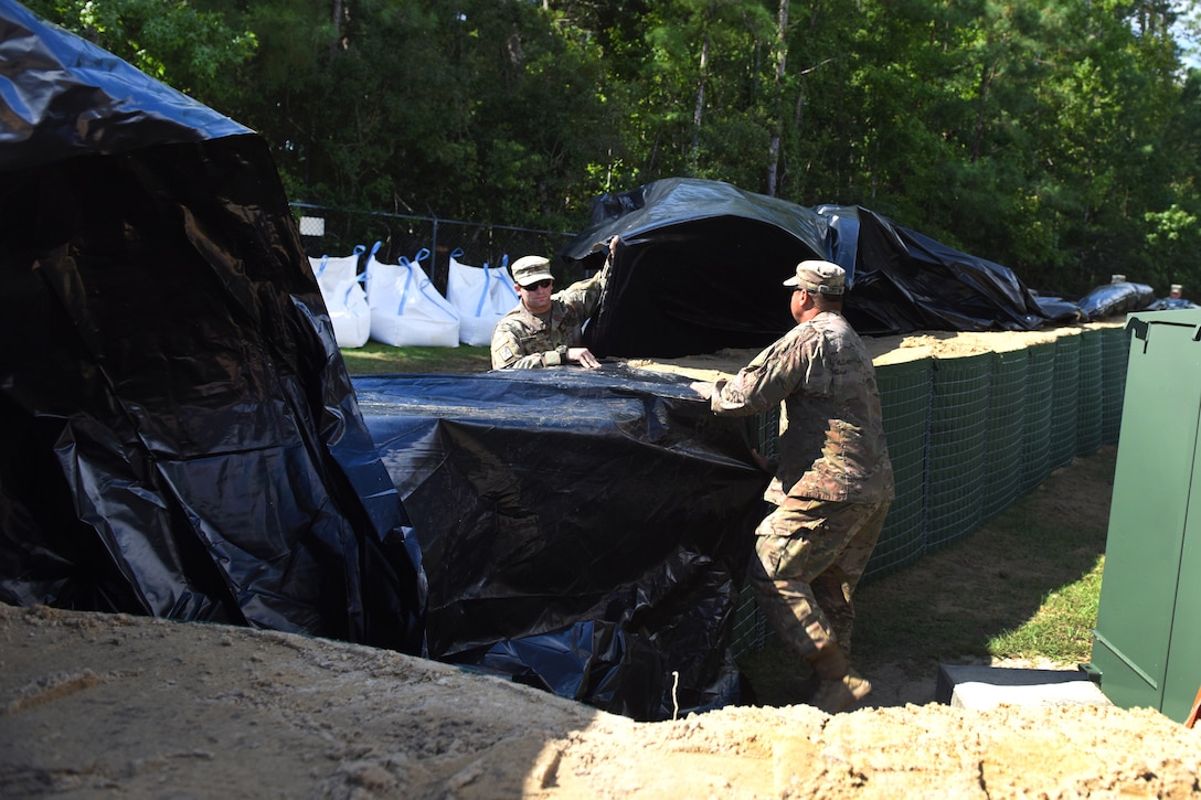 Soldier use sheets of plastic to cover flood water barriers filled with sand.