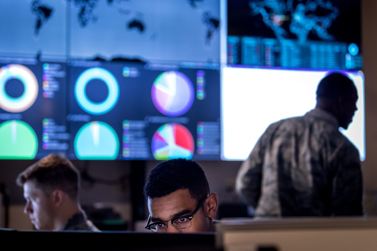 A man stands in front of a computer screen while other men are seated at desks in front of computer screens