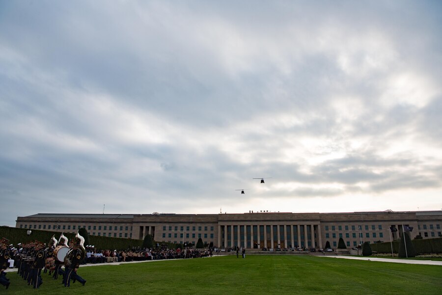 Two helicopters fly over the Pentagon as military musicians march on the parade field.