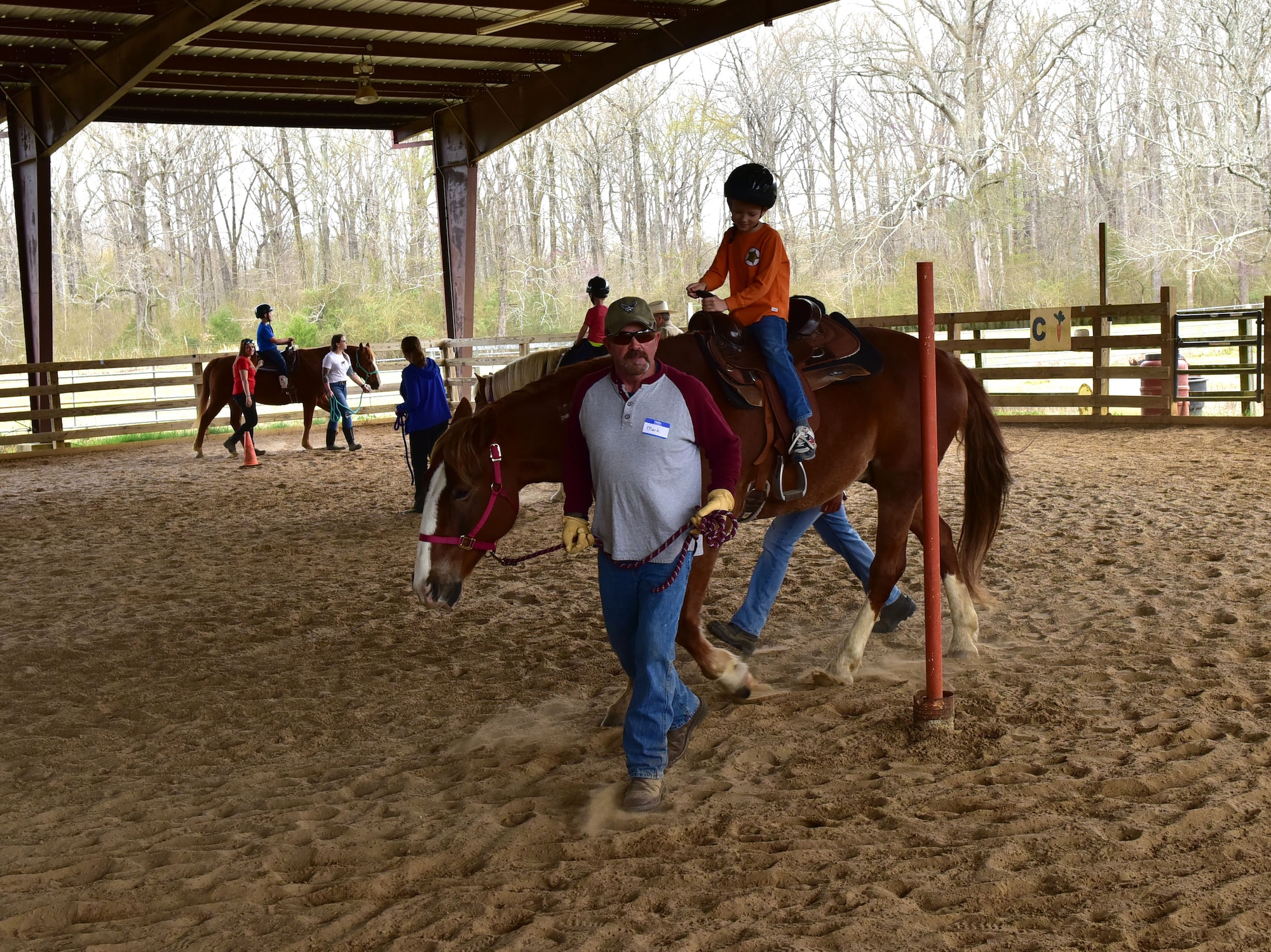 Male and female adults lead children on horseback around a dirt riding ring enclosed with a wooden fence.