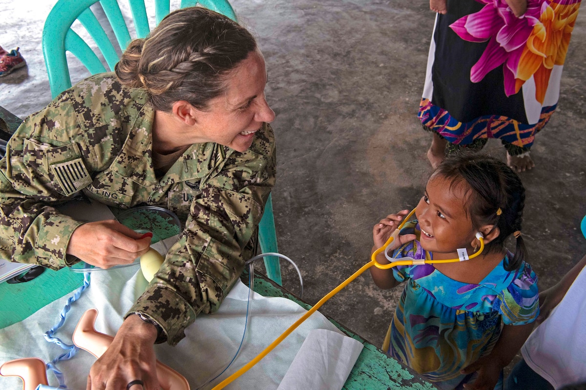 A sailor smiles while looking at a child wearing a stethoscope .
