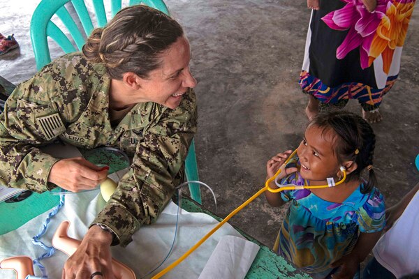 A sailor smiles while looking at a child wearing a stethoscope .