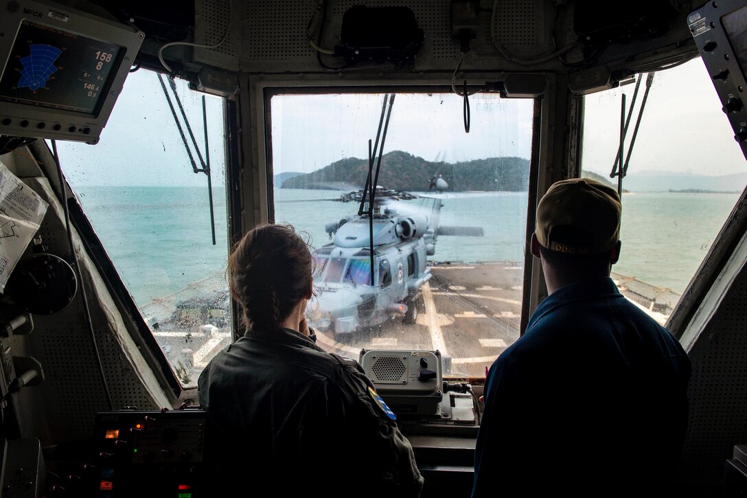 Two sailors look out a window towards a parked helicopter.