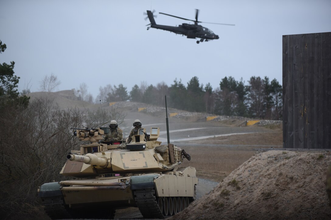 A tank drives as a helicopter flies above.