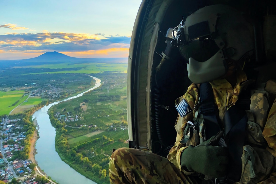 A soldier in a flight helmet looks out from an open helicopter over a river.