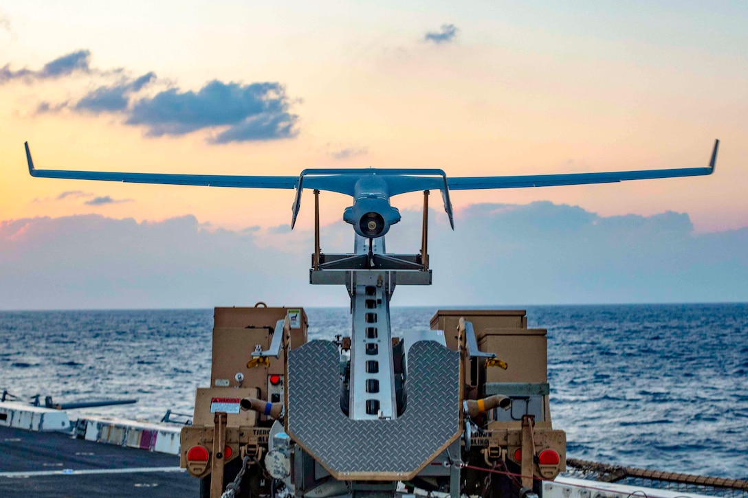 A drone sits on a launching device on the flight deck of a ship in water.