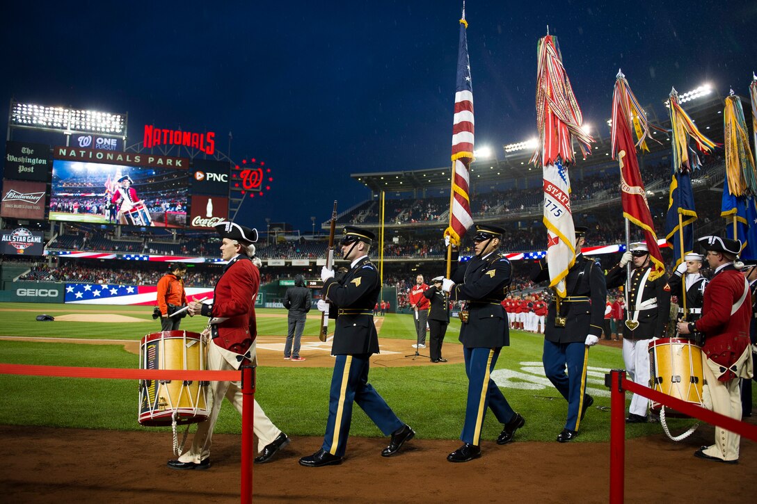 A line of service members carrying flags and playing a drum walk in a line at a baseball game.