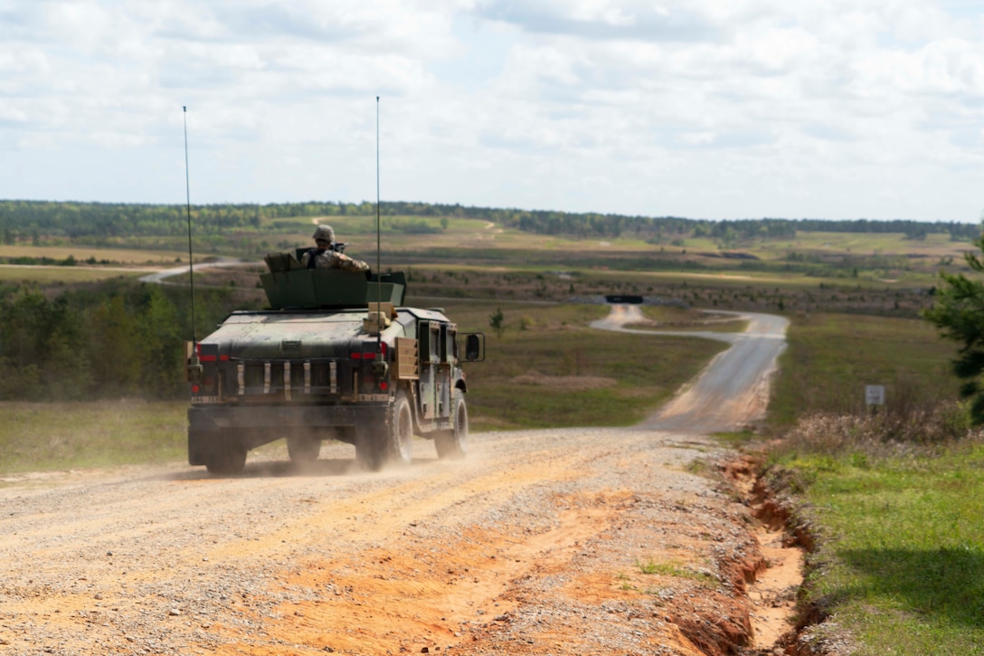 A service member sits in a military vehicle looking out over a flat field with a long dirt road.