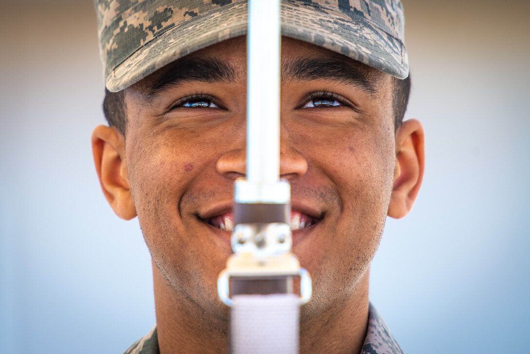 An Air Force honor guard member, shown in closeup, smiles while holding a weapon in front of the center of his face.