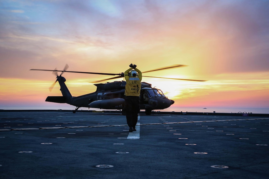 A sailor signals an aircrew in a helicopter on a military ship at twilight.