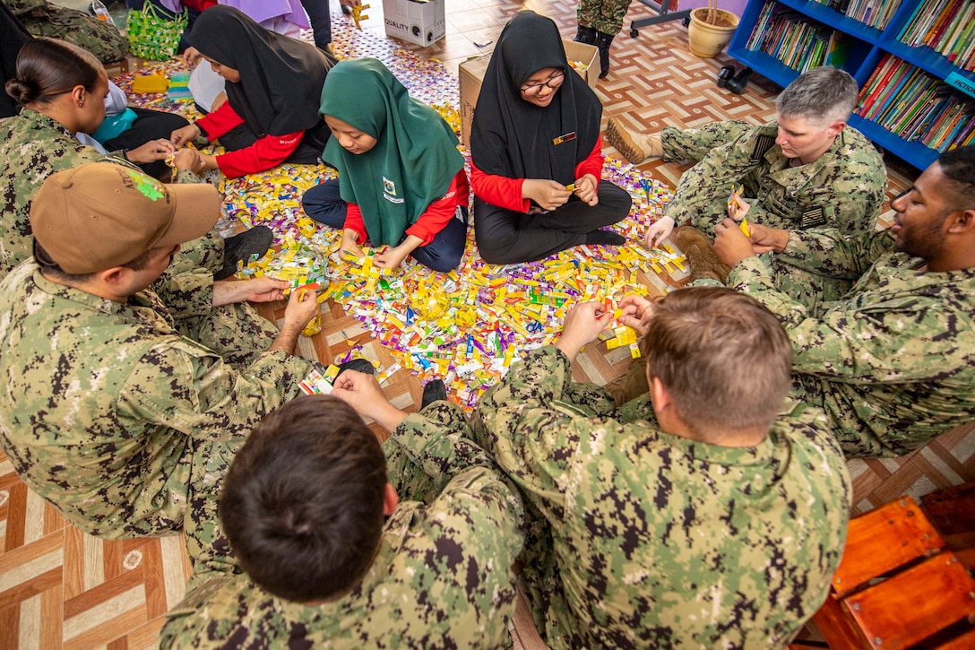 A group of sailors sit in a half circle in  front of three students with cardboard pieces between.