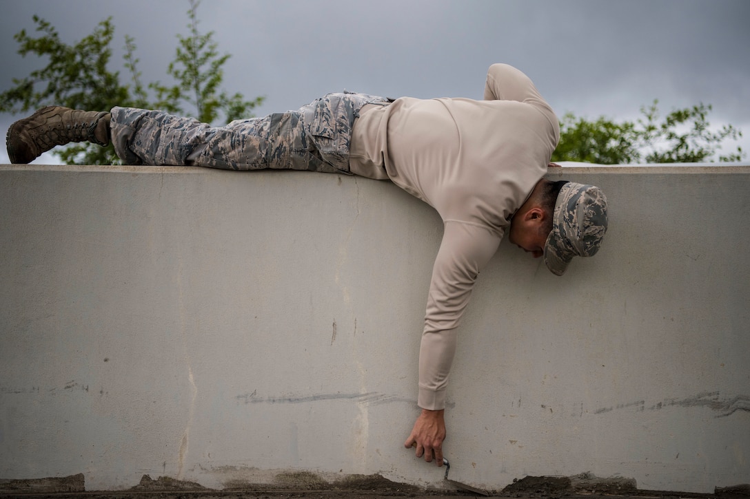 An airman lies on top of a concrete wall while using a tool to even out wet concrete on the floor.