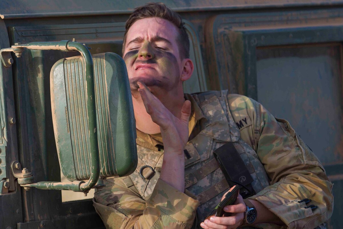 A soldier looks into the side mirror of a vehicle while applying paint to his face.