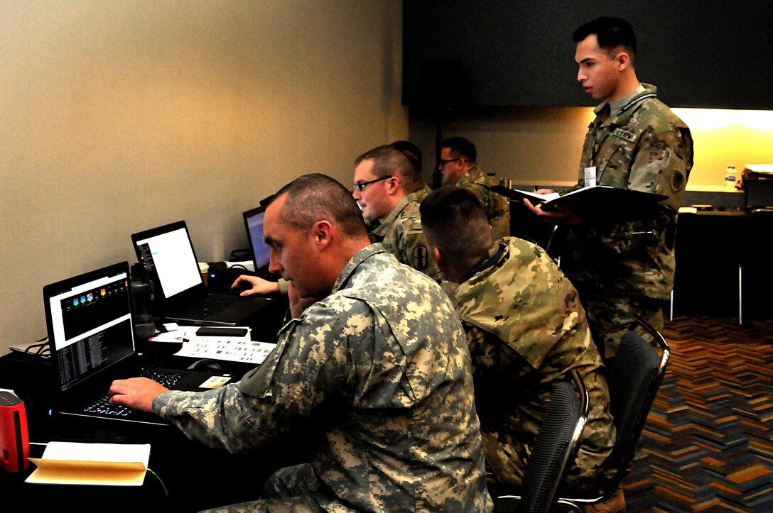 A soldier looks on as other troops type into laptops.
