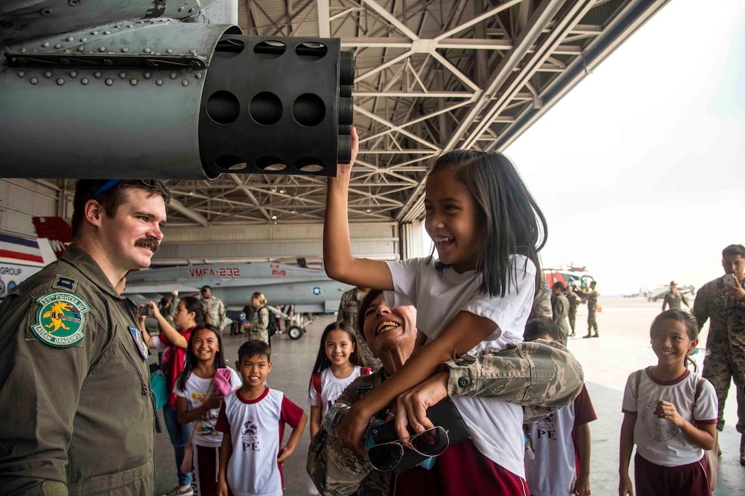 An airman lifts up a child who is touching part of an aircraft while a group of people surround the two.