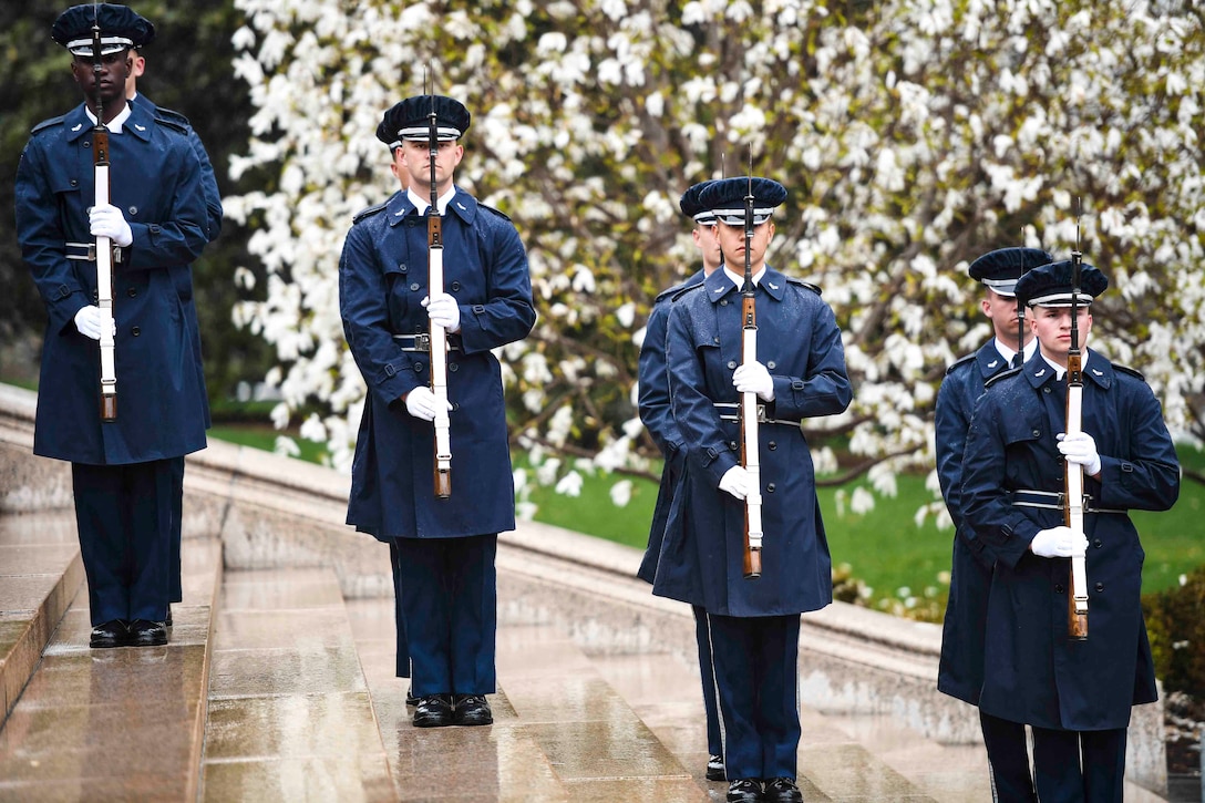 Eight airmen stand in two rows along steps while holding weapons at their chests.