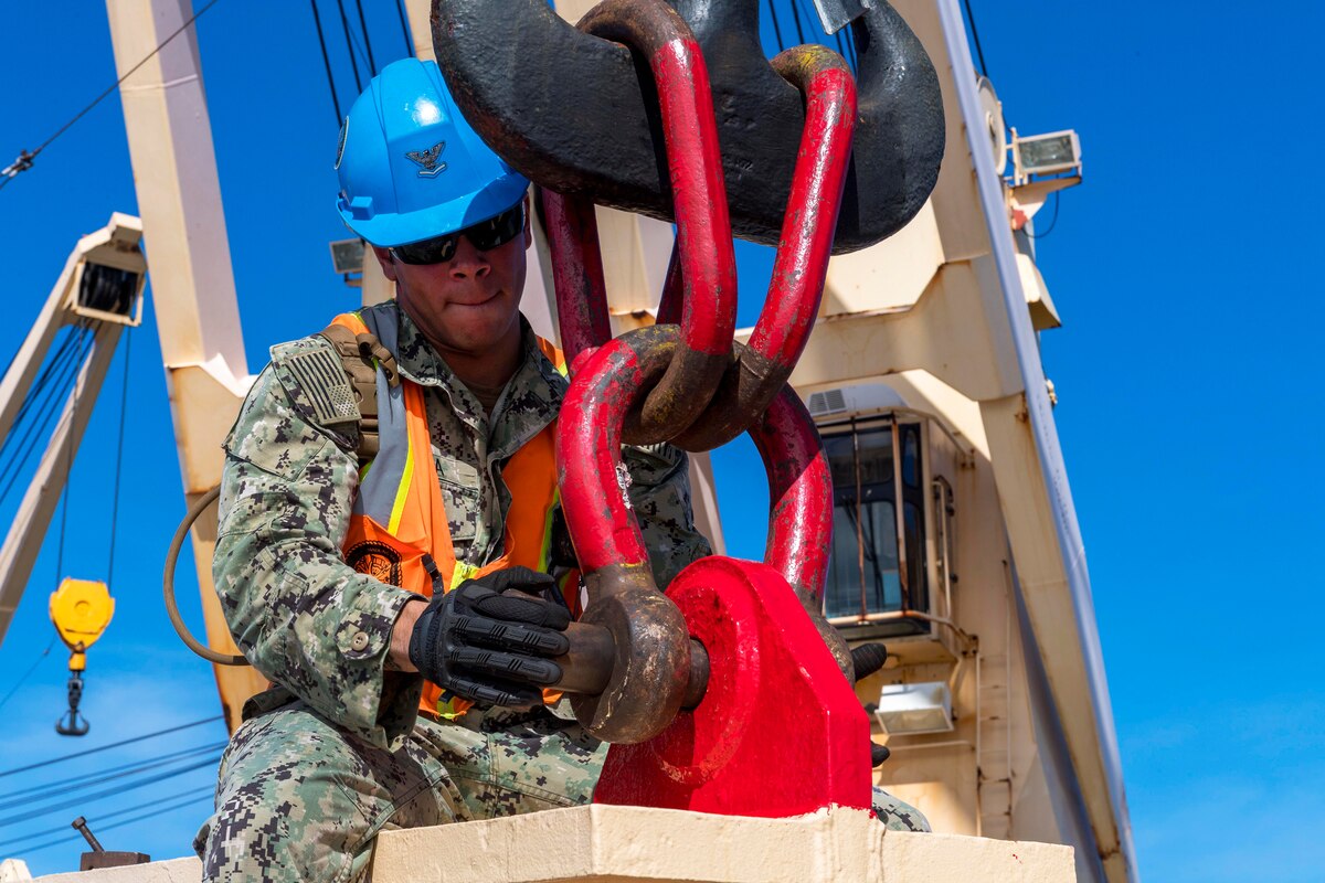 A sailor holds on to a large red hook as he attaches it to a beam.