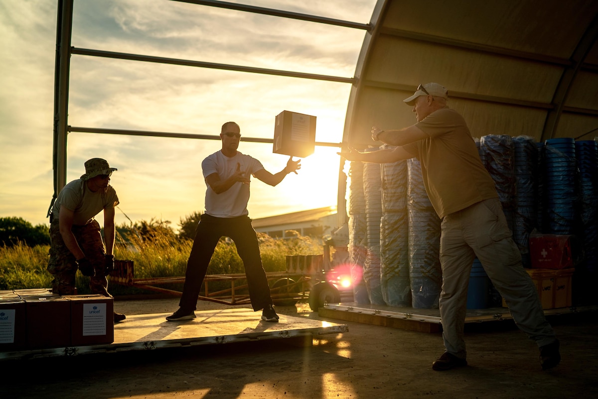 Three men throw boxes of supplies to load on a pallet at twilight.