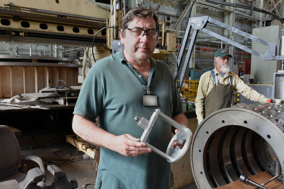 James “Trapper” Landon, an inside machinist in the Arnold Air Force Base Model and Machine Shop, left, shows one of the cages he constructed for the Trapper Lapper 5000, a device developed to speed the lapping process. In the background, Outside Machinist Joel Sizemore puts the Trapper Lapper 5000 to work. The device allows for the automation of a process that previously had to be performed manually. (U.S. Air Force photo by Bradley Hicks) (This image has been altered by obscuring a badge for security purposes)