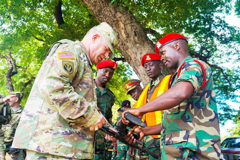 A soldier looks down at a gun he and a foreign soldier are both holding as other soldiers watch.