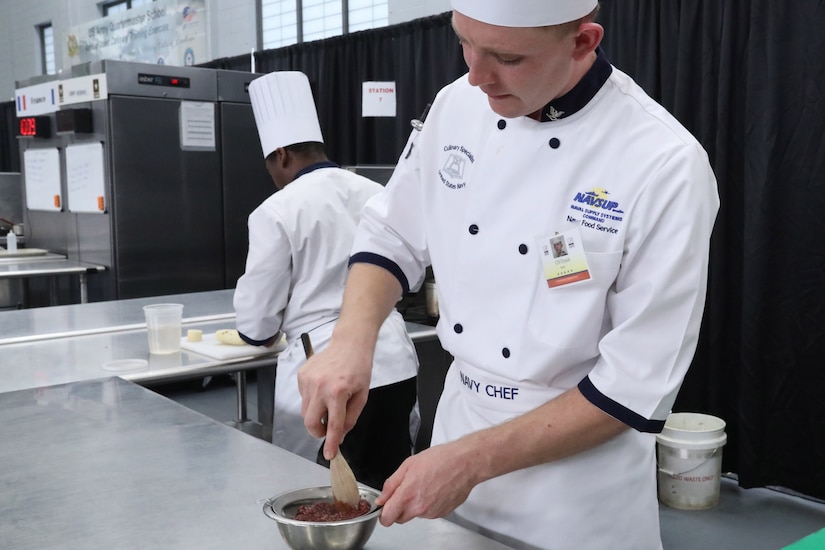 A chef pushes berries through a strainer while another chef works behind him.