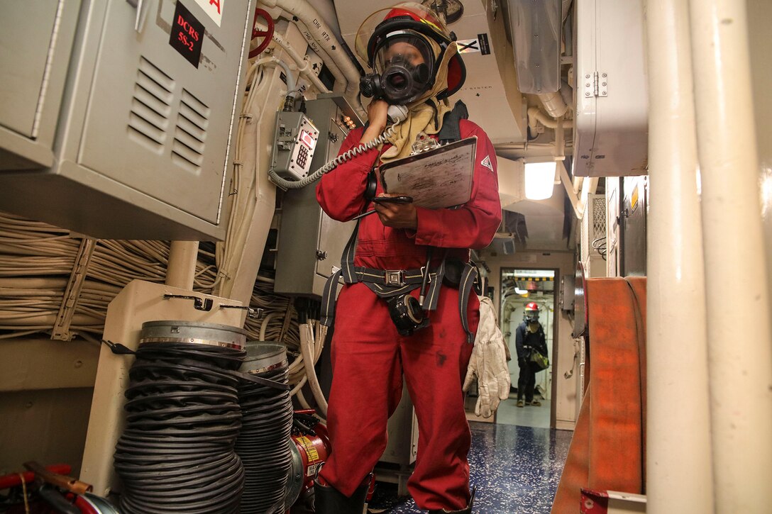 A sailor dressed in firefighting gear communicates via radio aboard a ship.