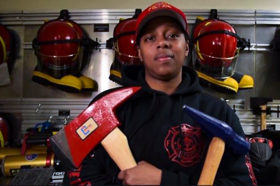 A sailor poses for a photo holding an ax and a hammer while standing in front of a wall of hanging firefighter helmets and boots.