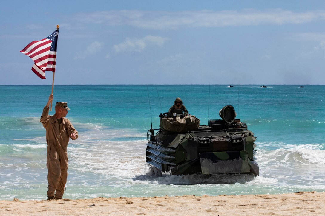 A Marine waves a flag while standing on a beach as an assault vehicle comes ashore.