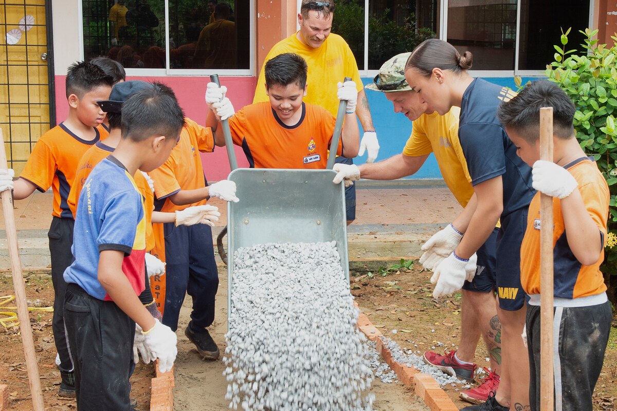 A student pours a wheelbarrow of stones for a walkway as other students and adults look on.