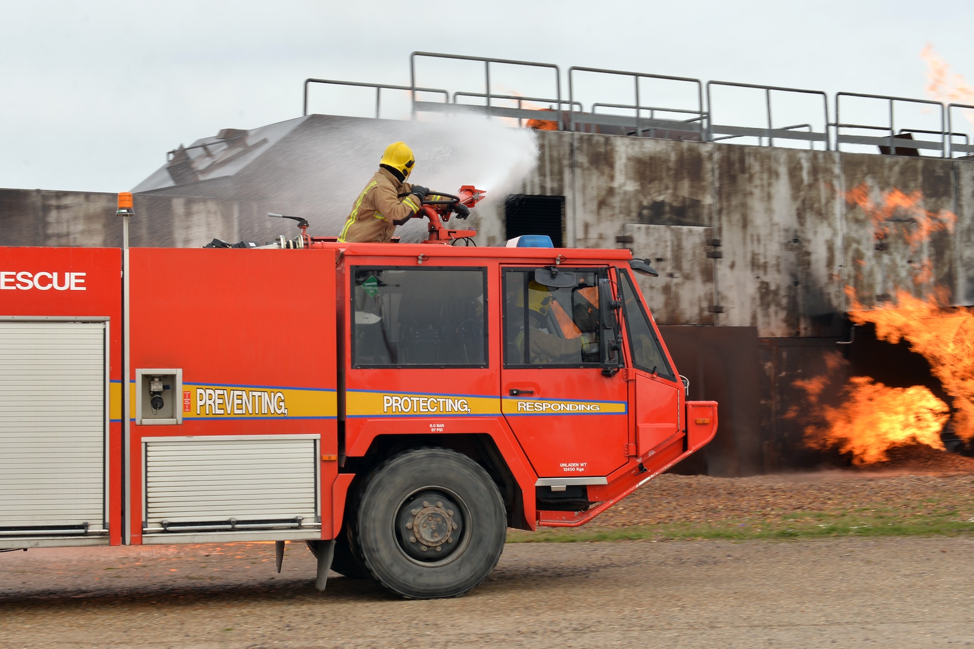 A firefighter from RAF Marham sprays water from a Royal Air Force fire truck onto a mock aircraft fire at the live fire training area on RAF Mildenhall, England, April 5, 2019. The British fire crew visited the base to gain experience on the live fire trainer. With the arrival of the F-35 and the regeneration at RAF Marham, their training facilities are under renovation and the visit served to provide vital training for the RAF firefighters, and aid in continuing to build vital working relationships between RAF Mildenhall and neighboring fire departments. (U.S. Air Force photo by Karen Abeyasekere)