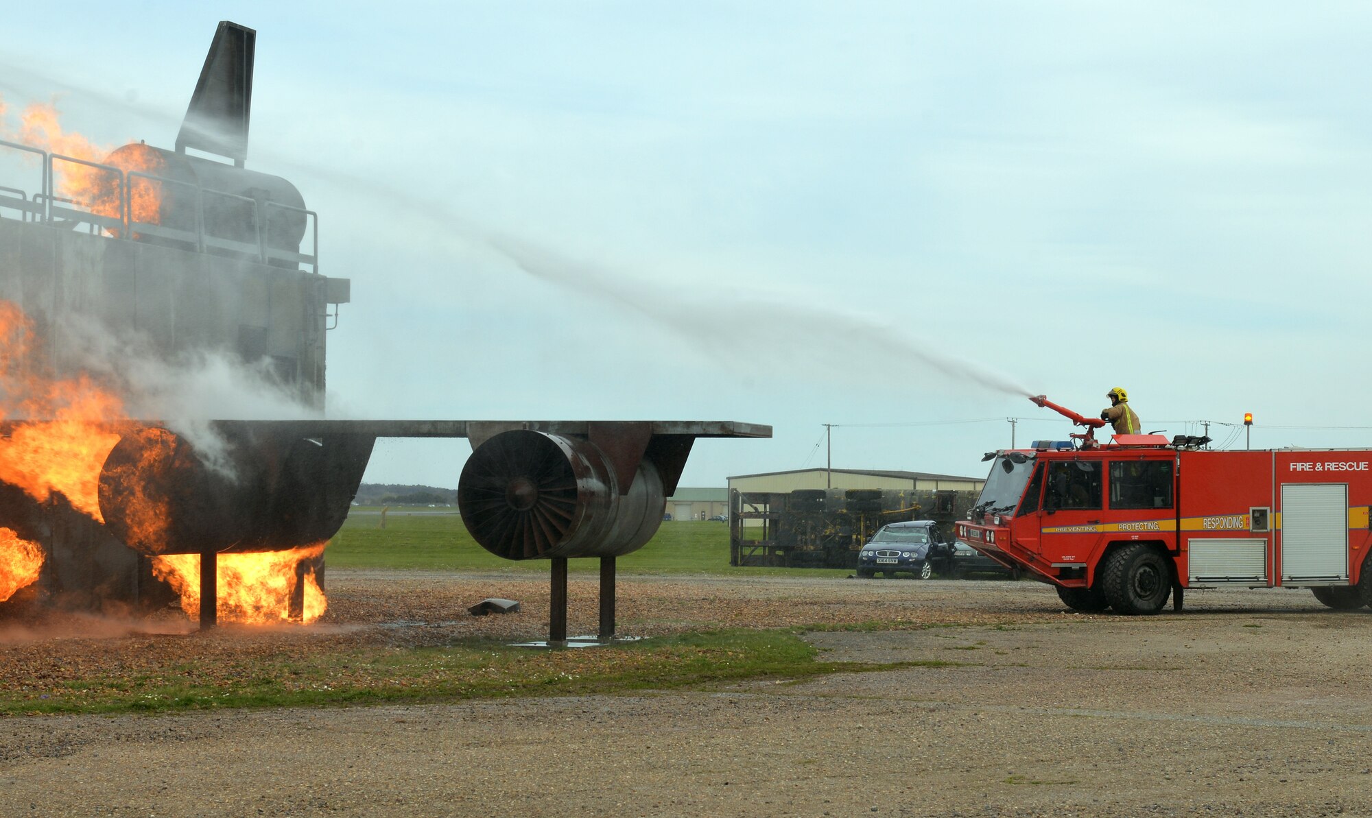 A firefighter from RAF Marham sprays water from a Royal Air Force fire truck onto a mock aircraft fire at the live fire training area on RAF Mildenhall, England, April 5, 2019. The British fire crew visited the base to gain experience on the live fire trainer. With the arrival of the F-35 and the regeneration at RAF Marham, their training facilities are under renovation and the visit served to provide vital training for the RAF firefighters and aid in continuing to build vital working relationships between RAF Mildenhall and neighboring fire departments. (U.S. Air Force photo by Karen Abeyasekere)