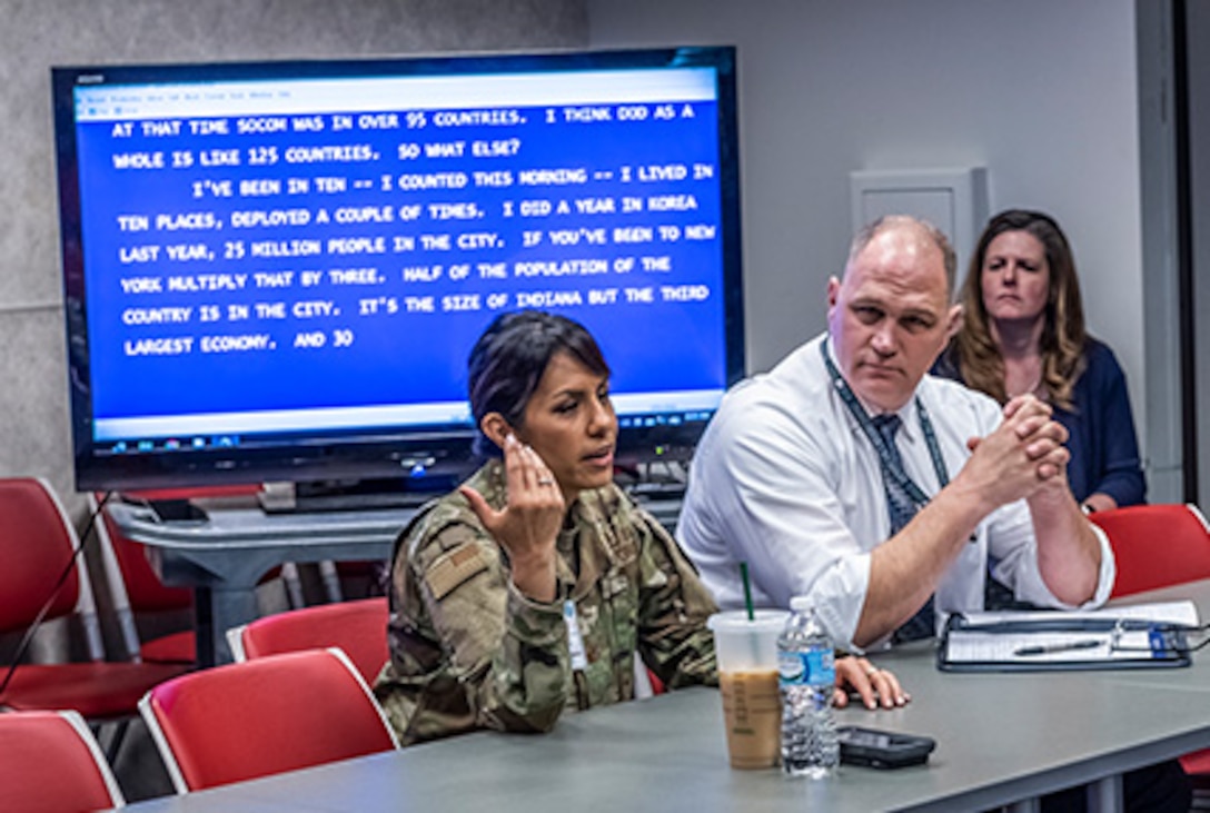 Military woman sits next to civilian man both in front of a blue screen tv with text