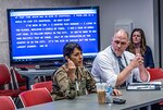 Military woman sits next to civilian man both in front of a blue screen tv with text