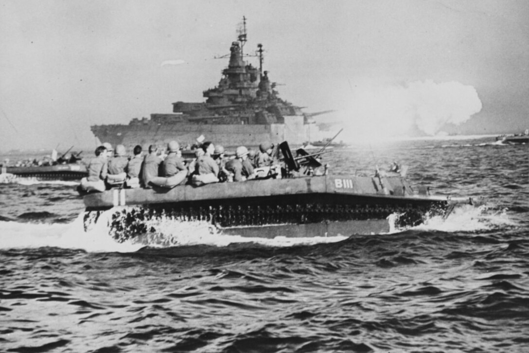 Two landing crafts filled with Marines cut through water as a ship in the distance fires its gun.