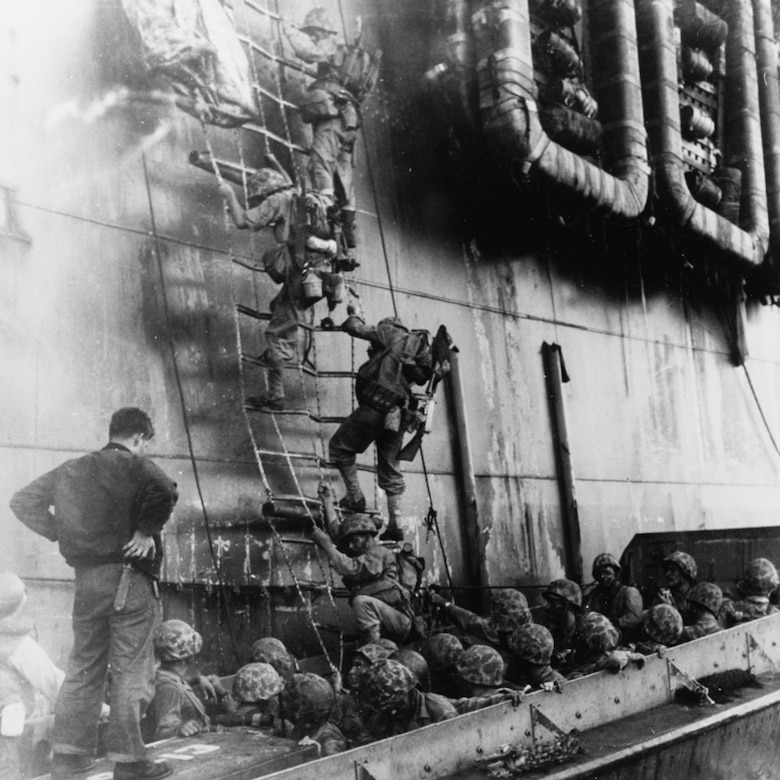 Marines climb down a net on the side of a ship into a small boat filled with more Marines.