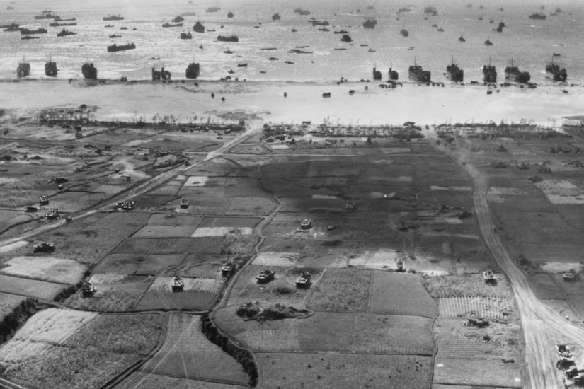 An aerial view of a beach with tracked vehicles on land and ships in the water.