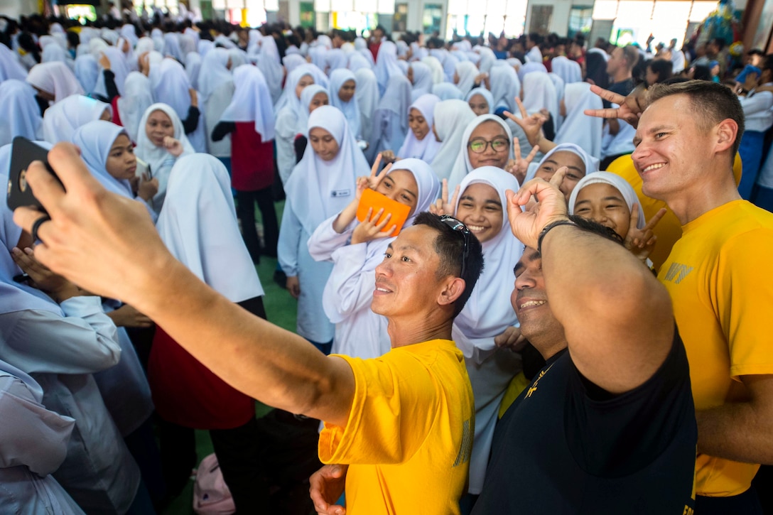 Three sailors take a selfie with a large group of students.