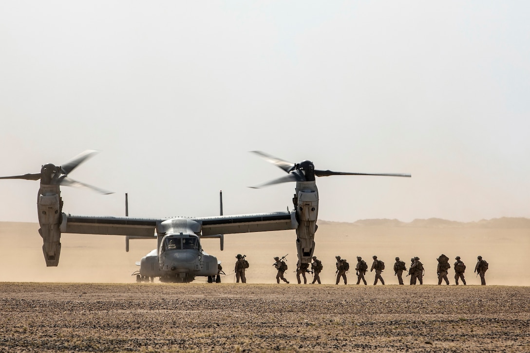 Marines and sailors walk in a line on desert terrain to a parked Osprey.