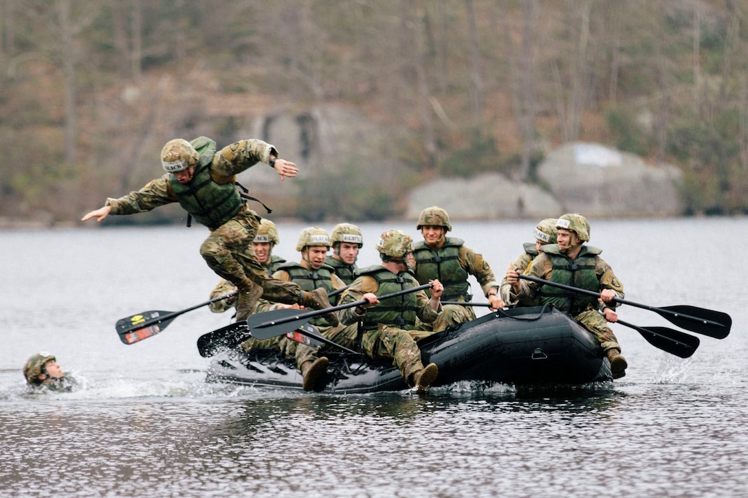 A group of soldiers paddle through water in a rubber boat as one soldier jumps into the water.