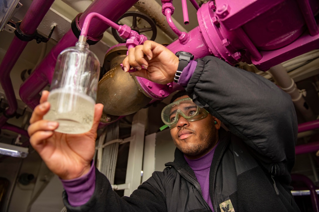 A service member holds a small jar as a liquid  drips into it from a pipe above him.