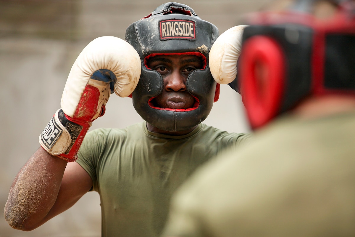 A Marine Corps recruit wearing protective headgear and boxing gloves holds up his fists while facing an opponent.