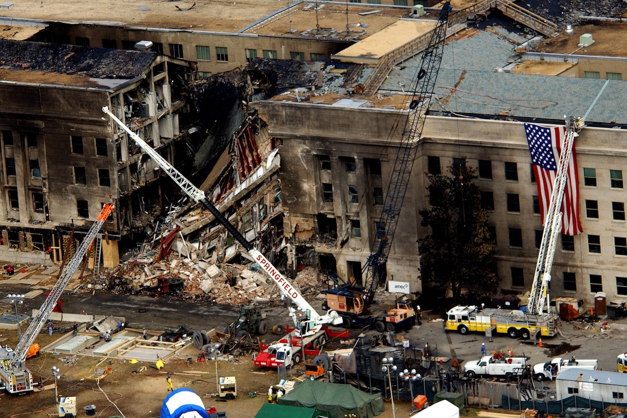 An aerial view of the charred and partially collapsed Pentagon, with fire trucks and emergency crews working around rubble.