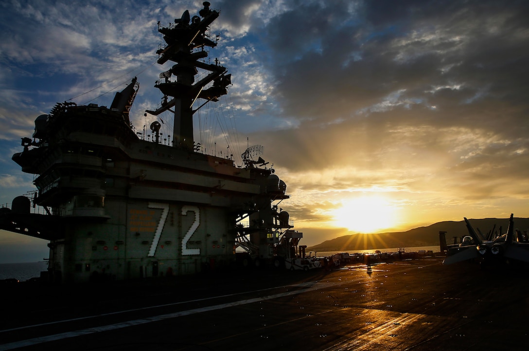 The sun sets behind an anchored aircraft carrier.