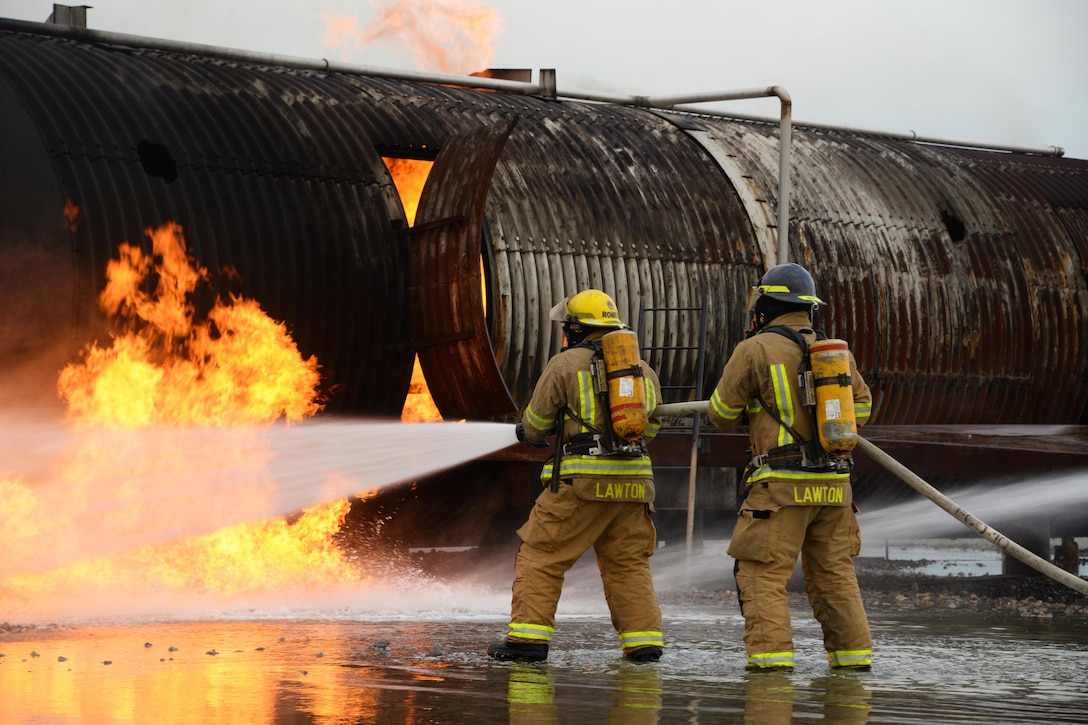 Two firefighters point a  water hose toward a burning fuselage during training.
