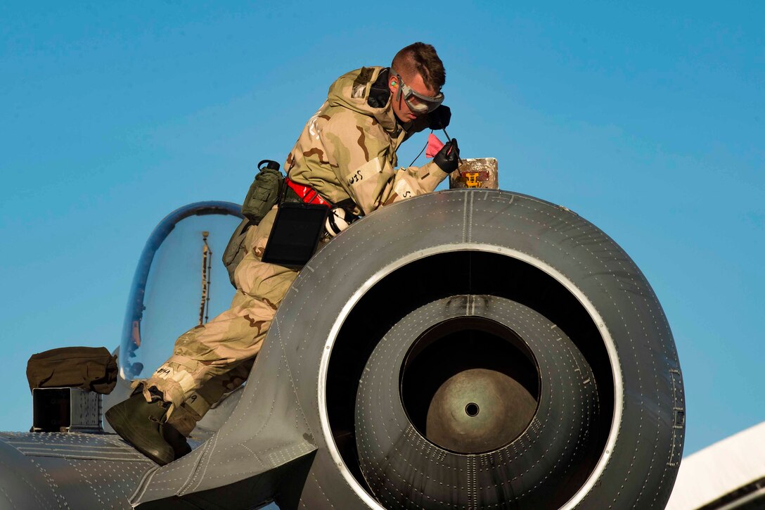 An airman lies on top of an aircraft holding an oil stick towards an opening.