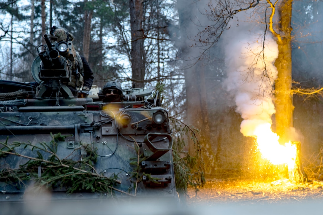 A tank rolls toward the camera as a tree burns in the background.