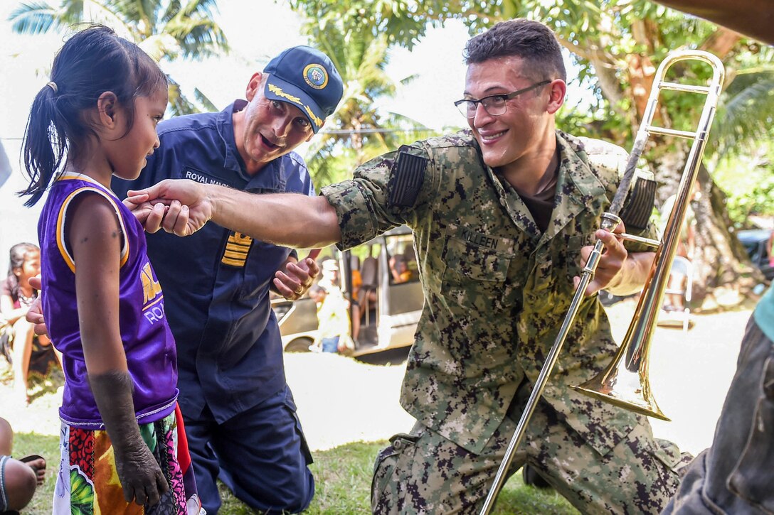 A sailor with a trombone smiles and holds his hand under a girl's chin as another service member stands by.