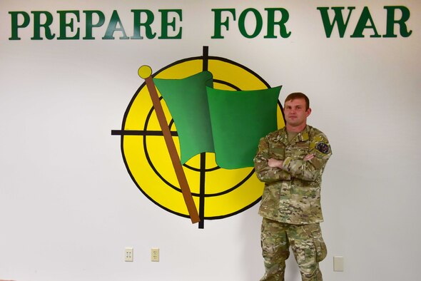 A man wearing the operational camouflage pattern uniform stand with his arms folded in front of a mural of a gren flag.