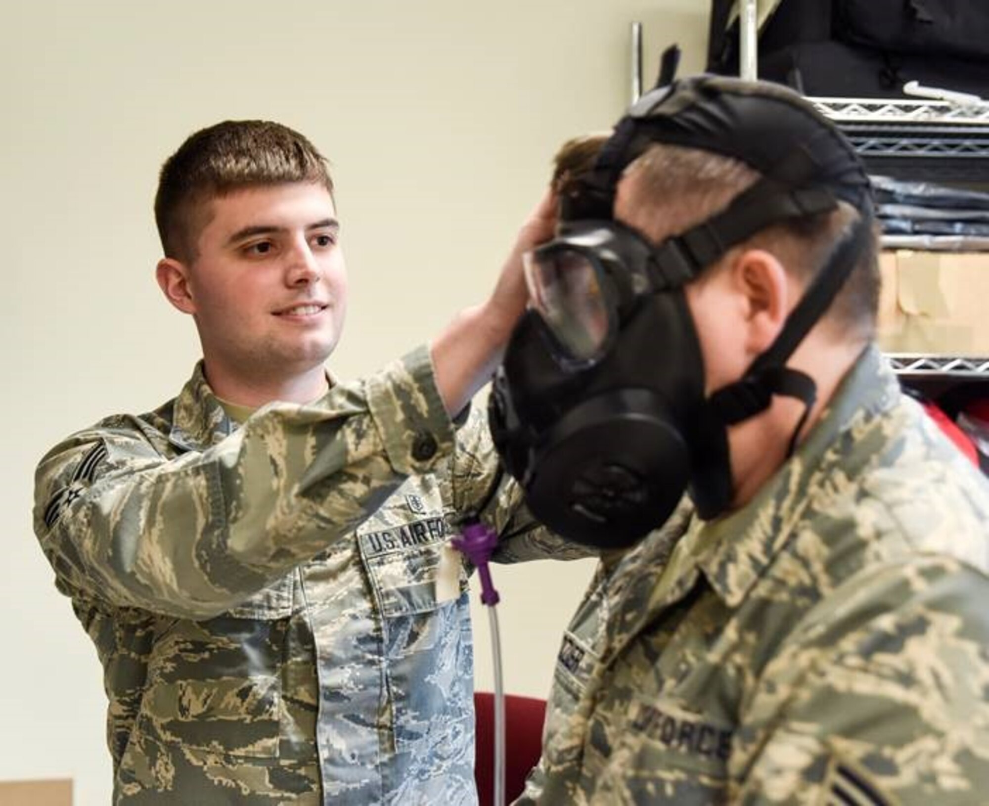 A man wearing the Airman Battle Uniform fits a gas mask on another man wearing the Airman Battle Uniform.